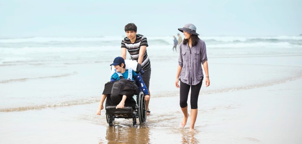 Older brother and sister walking younger disabled brotherin wheelchair in the water along the beach at ocean in summer time