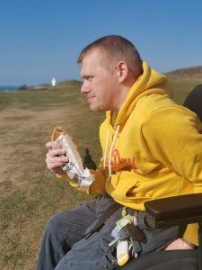 Nick Carr with a yellow jacket on, eating a pasty, looking out to sea