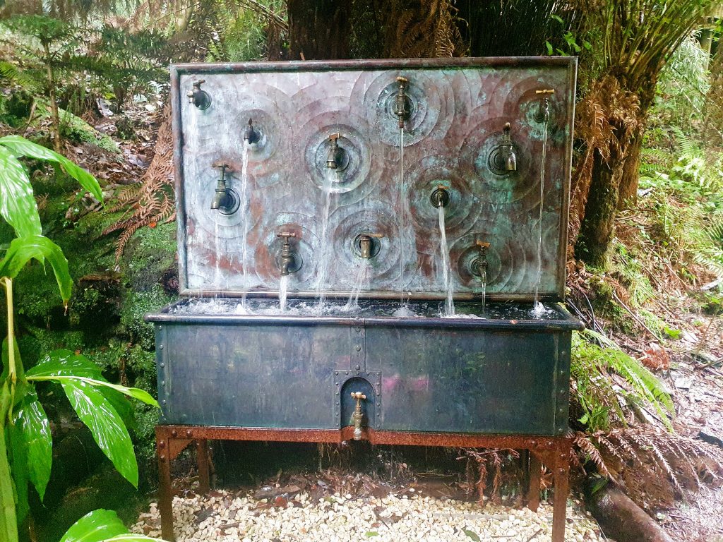 A sculpture comprising of a large metal sink with a backboard. On the backboard are lots of taps with water flowing into the sink below