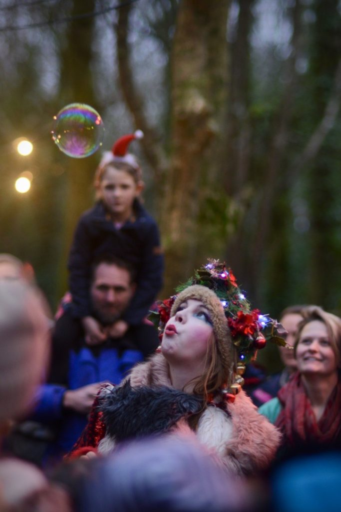 A performer in the woods blowing bubbles with the audience, including a father with a child on his shoulders looking on, surrounded by woodland and fairy lights