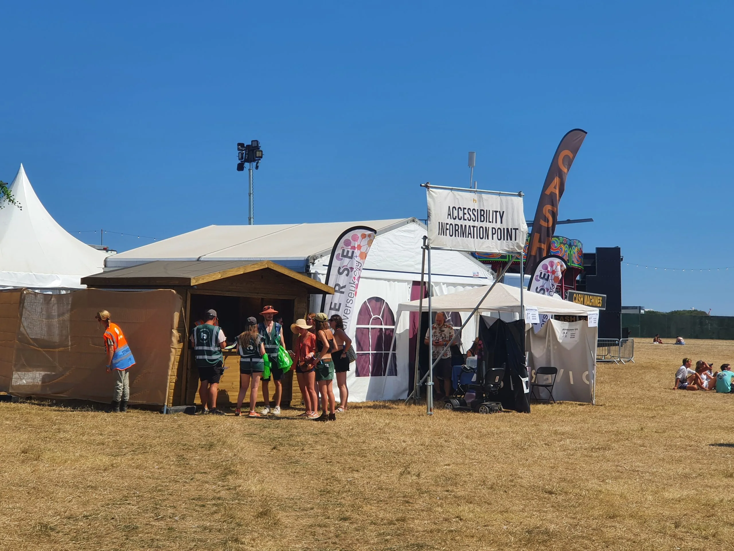 Accessibility information point tent along with a cash point sign. 3 stewards can be seen in the front of the image wearing bright green vests 