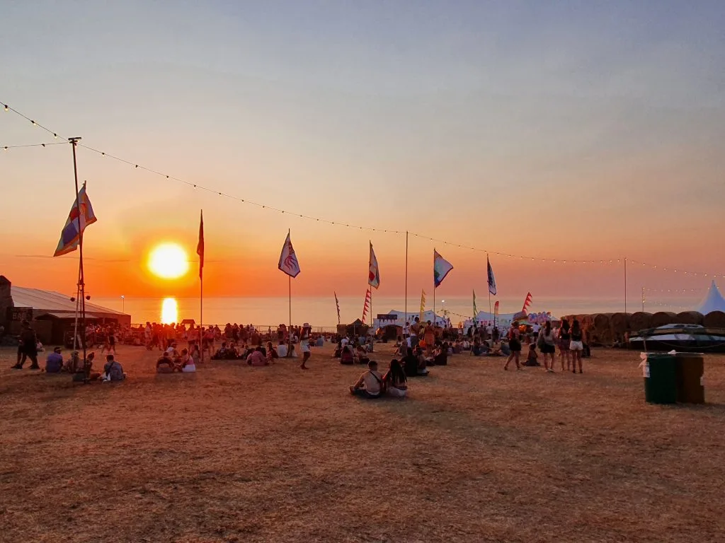 Boardmasters festival at sunset, the sun is setting over the ocean and people are sitting in groups under flags on the beach.