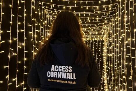 Viki at Access Cornwall wearing a black hoodie with the Access Cornwall name standings looking into a tunnel of golden lights at the Tunnel of lights Charlestown Shipwreck Museum, St Austell