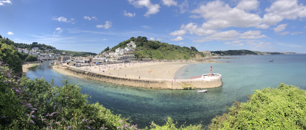 panoramic view of the river flowing out of Looe, from the left of the photo, past the pier, with the beach protected behind it, and hills behind. Lovely blue water.