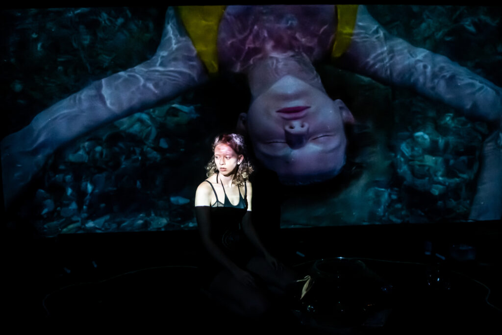 a young woman looking pensive, standing in front of a projected image of herself on a large screen. in the projected image she is seen with her eyes closed with water patterns on her arms 