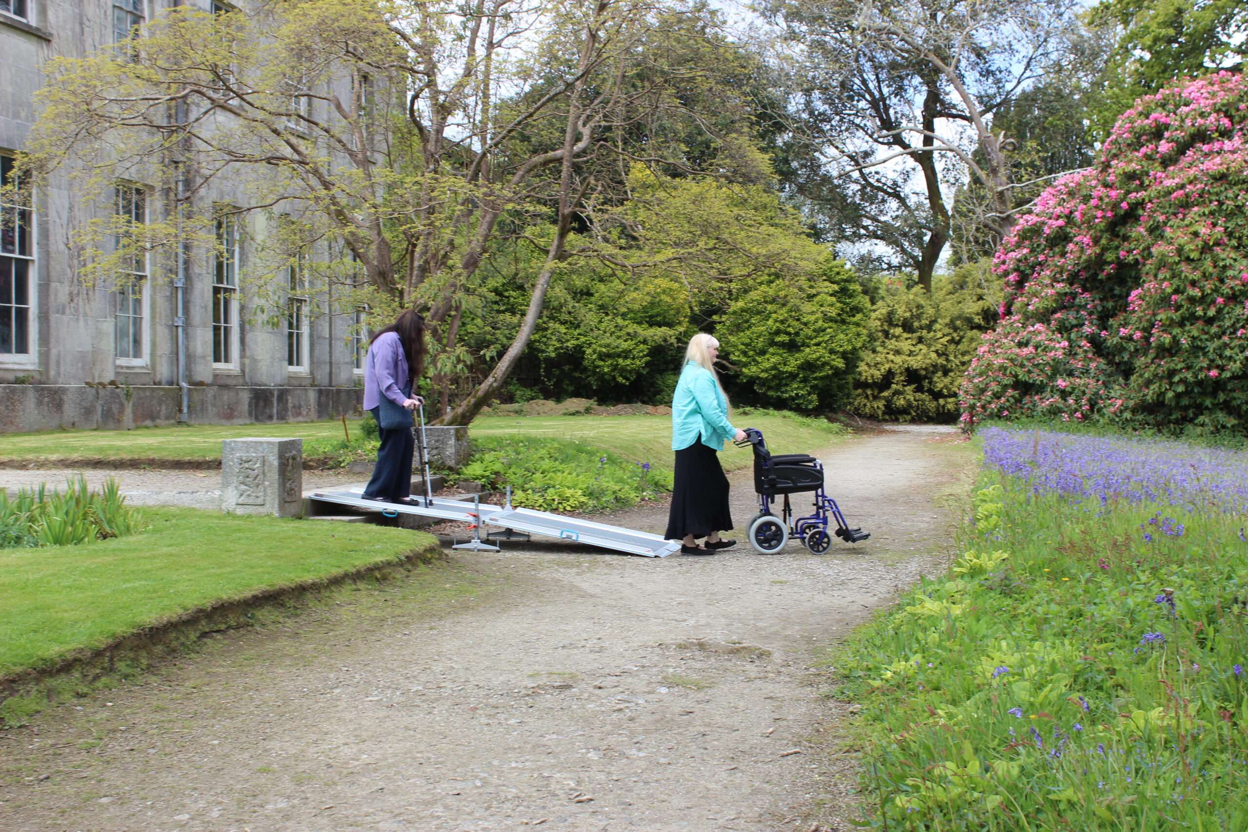 A lady pushing a wheelchair and a lady walking down a ramp using her walking aid at Enys Gardens
