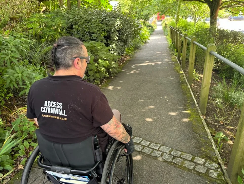 An Access Cornwall auditor at work. The auditor is a white man using his wheelchair and he has the Access Cornwall name in white on the back of his black shirt