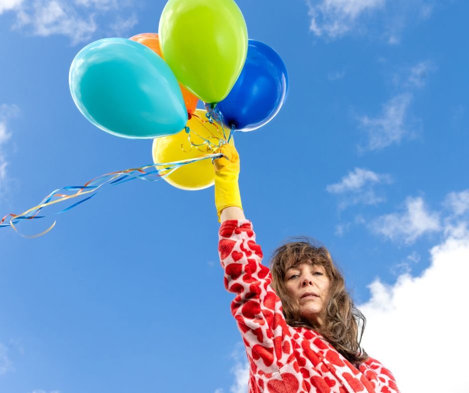 A woman in a red dress holding balloons against a blue sky. The image promoting the play SCAFFOLDING