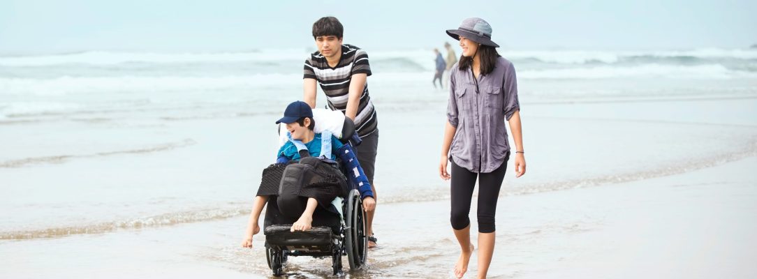 Older brother and sister walking younger disabled brotherin wheelchair in the water along the beach at ocean in summer time